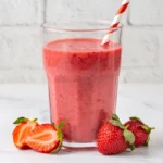 Pink strawberry smoothie in a tall glass with a red-and-white striped straw, surrounded by fresh strawberries on a light marble surface.