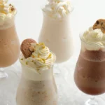 Assorted dessert drinks topped with whipped cream in clear glasses with cookies nearby on a bright surface.