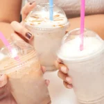 Close-up of hands holding three iced milkshakes topped with whipped cream and caramel drizzle, with pink and blue straws in clear cups.