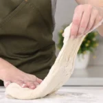 Close-up of hands kneading soft dough on a floured surface, wearing a green apron.