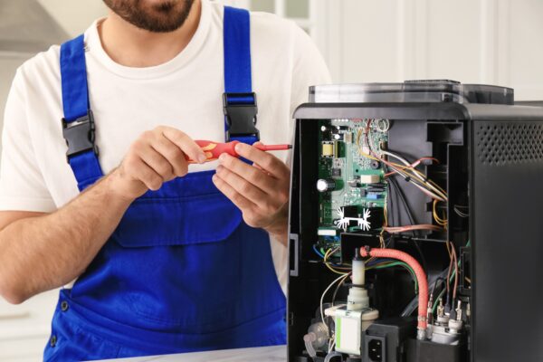 Technician in blue overalls using a screwdriver to repair a printer with exposed circuitry.